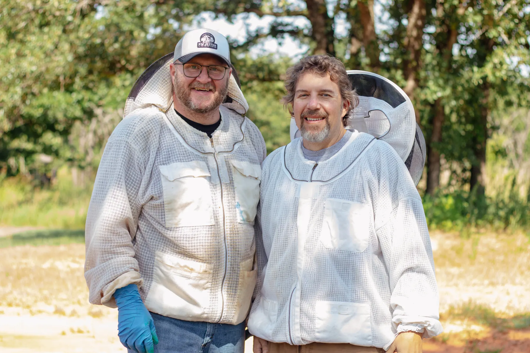 Jimmy Hughes and Jimmy Neal of Oklahoma Hive Patrol in white beekeeping jackets, smiling outdoors.