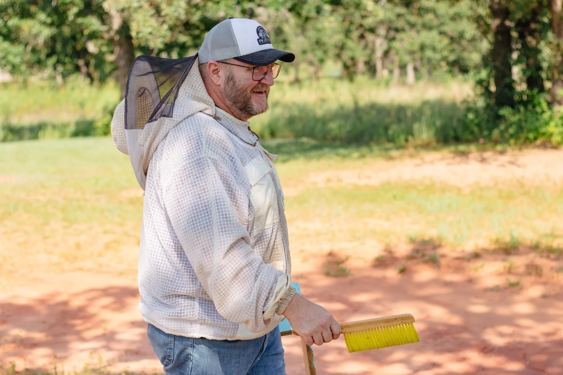 Jimmy Neal in a ventilated beekeeping jacket smiling outdoors, holding a yellow bee brush.