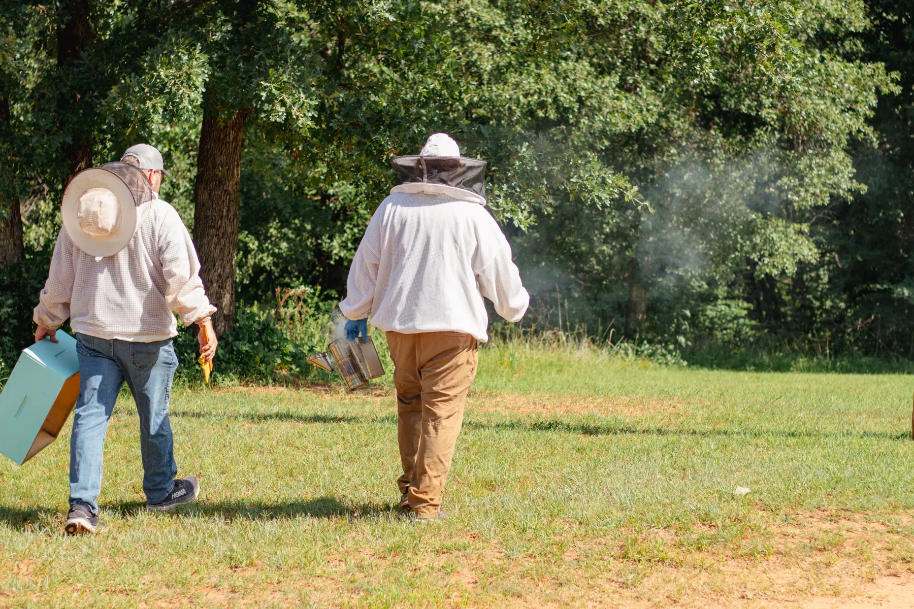 Oklahoma Hive Patrol walking across a field toward the trees, one carrying a hive box and the other a smoker.